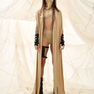 May include: A tan hooded cape and matching outfit. The outfit includes a top, shorts, and a face covering. Black arm cuffs and leg straps add detail. The model is wearing black platform boots. The background is a light fabric.