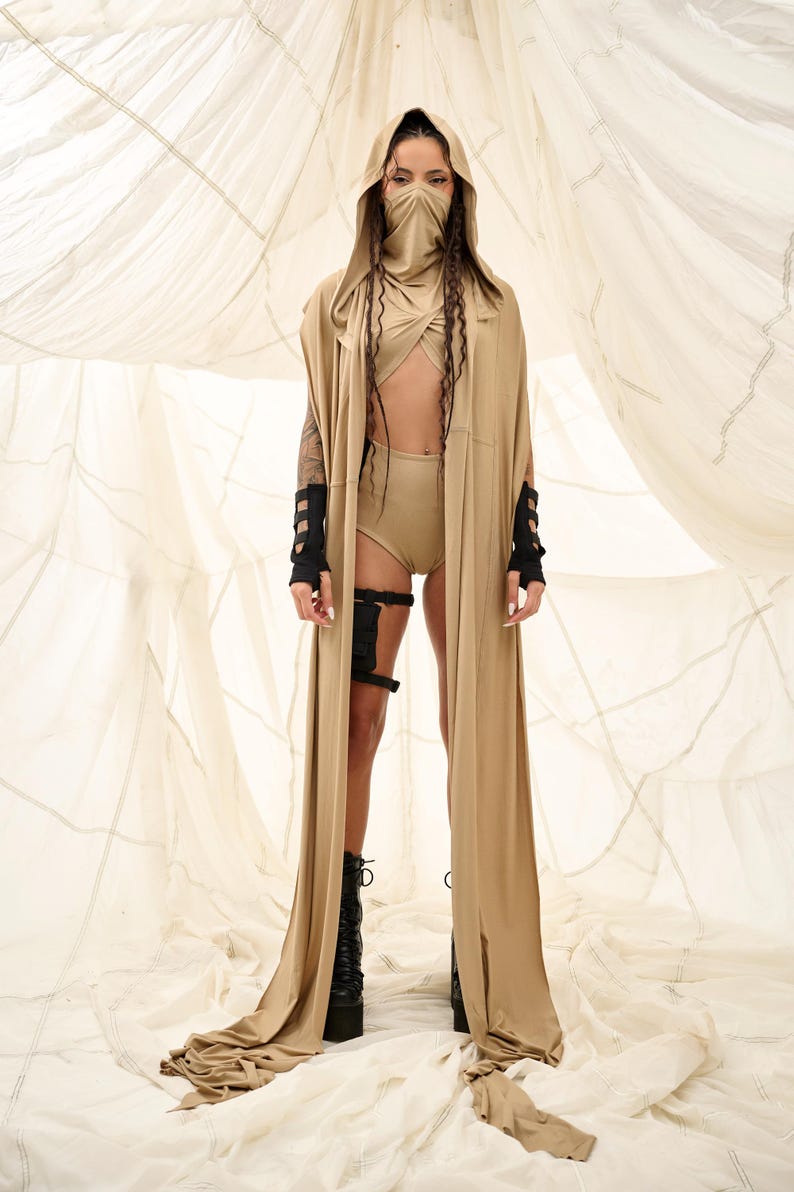 May include: A tan hooded cape and matching outfit. The outfit includes a top, shorts, and a face covering. Black arm cuffs and leg straps add detail. The model is wearing black platform boots. The background is a light fabric.
