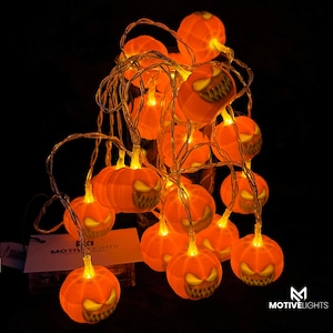 Könnte beinhalten: Lichterkette mit orangefarbenen Kürbissen mit geschnitzten, grinsenden Gesichtern. Die Halloween-Lichter sind beleuchtet und werfen einen warmen Schein auf einen dunklen Hintergrund. Die Lichter sind auf einem dünnen, transparenten Draht aufgereiht.