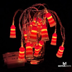 Könnte beinhalten: Lichterkette mit Miniatur-Leuchtturm-Designs mit roten, gelben und weißen Streifen. Die Lichter sind auf einem dünnen Draht aufgereiht und leuchten. Ein kleiner weißer Akku ist sichtbar.