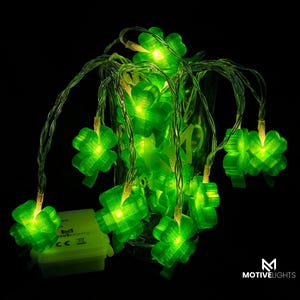 Könnte beinhalten: Eine Lichterkette mit grünen, leuchtenden LED-Klee-förmigen Lichtern. Die Lichter sind in einem Glasbehälter vor schwarzem Hintergrund angeordnet. Ein kleiner, grüner Akku ist sichtbar. Der Markenname "MOTIVELIGHTS" wird angezeigt.