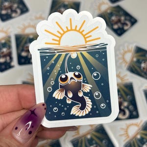 May include: A sticker featuring a cartoon anglerfish in a dark blue underwater scene with bubbles. The sticker has a white border and a sun design at the top. The anglerfish is black with white and tan fins and large eyes.