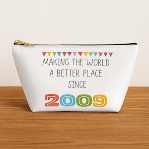 May include: White canvas cosmetic bag with a black zipper and gold pull. The bag features the text "MAKING THE WORLD A BETTER PLACE SINCE 2009" with colorful banner accents. The bag is on a wooden surface.