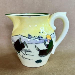 May include: Small, vintage ceramic pitcher with a yellow body and a green rim. The pitcher features a hand-painted scene with figures, mountains, and a bird. The handle is white, and the overall design is reminiscent of antique pottery.