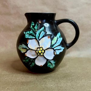 Puede incluir: Una pequeña jarra de cerámica negra con un diseño de flor blanca. La flor tiene un centro amarillo y está rodeada de hojas verdes y azules. La jarra tiene un cuerpo redondeado, un asa y un pico.