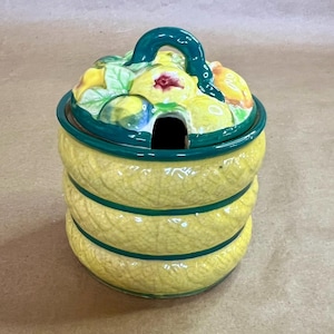 May include: A yellow ceramic jar with a green rim and handle. The jar has a textured, woven appearance and is decorated with a fruit and leaf design on the lid. The lid has a small opening for a spoon. The jar is likely for serving condiments.