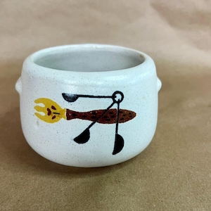 May include: A small, speckled white ceramic bowl with two small handles. The bowl features a whimsical illustration of a fork, spoon, and spatula in yellow, brown, and black. The bowl is likely for kitchen or dining use.