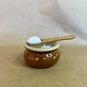 May include: A small, brown ceramic salt cellar with a wooden spoon resting on top. The spoon holds a small pile of white salt. The salt cellar is round with a slightly flared rim. The background is a solid, light brown color.