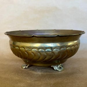 May include: A round brass bowl with a scalloped rim and a decorative embossed pattern of repeating curved shapes around the exterior. The bowl is supported by three small, ornate feet and features a polished metallic finish with visible patina.