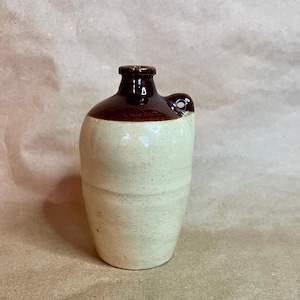 May include: A small, ceramic jug with a rounded body and a dark brown top and handle. The jug has a light beige speckled finish and a small opening. The handle is a loop shape. The jug is a decorative item.
