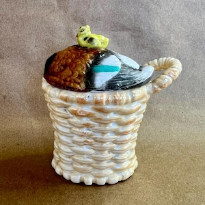 May include: A ceramic duck-shaped container with a woven basket design. The lid is brown, black, and teal, with a small yellow duck figurine on top. The container has a handle and is cream-colored.