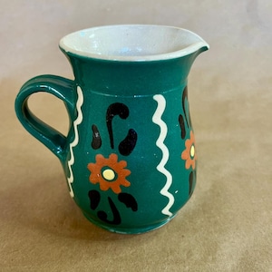 May include: A small, hand-painted ceramic pitcher in a vibrant teal color. The pitcher features a white rim and handle, with decorative orange and black floral designs and white wavy lines. This decorative pitcher is perfect for serving cream or sauces.