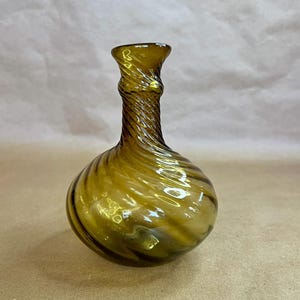 May include: A small, antique-style glass bottle with a bulbous body and a twisted neck. The bottle is a translucent amber color, with darker amber swirls throughout. The bottle is empty and sits on a neutral background.