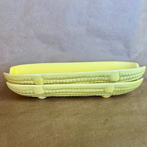 May include: Two-piece yellow ceramic butter dish shaped like corn on the cob. The dish has a textured surface mimicking corn kernels and a curved shape. The butter is a solid yellow color.
