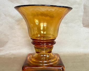 May include: Amber-colored glass vase with a flared top and a square base. The vase has a textured surface and a decorative design around the base. The glass has a warm, inviting hue, perfect for displaying flowers or as a decorative piece.