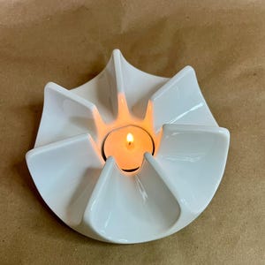 May include: A white ceramic candle holder with a flower-like design. The holder has six petal-shaped sections surrounding a central area where a lit tea light candle is placed. The candle flame emits a warm glow.