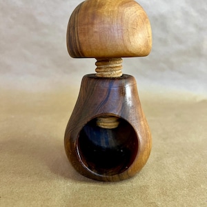 May include: A wooden nutcracker with a mushroom-shaped top and a pear-shaped base. The top is a lighter wood color, while the base is a darker, contrasting wood. The nutcracker has a screw mechanism for cracking nuts. The background is a neutral tan color.