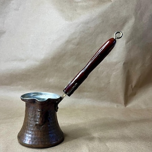 May include: A hammered copper coffee pot with a wooden handle. The pot has a traditional design and is perfect for brewing Turkish coffee.