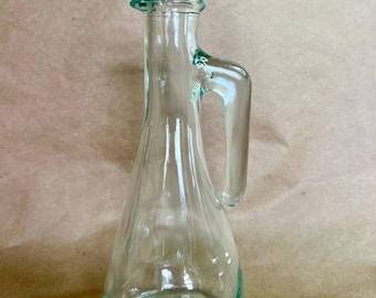 May include: Clear glass pitcher with a handle and a flared base. The pitcher has a rounded body and a small spout. The handle is attached to the side of the pitcher. The glass has a slight green tint. The pitcher is approximately 7 inches tall.