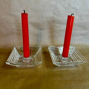 May include: Two clear glass square candle holders with red candles. The candle holders have a ribbed design and a circular base. The red candles are tall and cylindrical, with a black wick at the top. The background is a light brown.