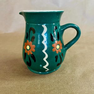 May include: A small, teal ceramic pitcher with a white rim and handle. The pitcher is decorated with orange and yellow floral designs and a white zigzag pattern. The pitcher is approximately 4 inches tall.