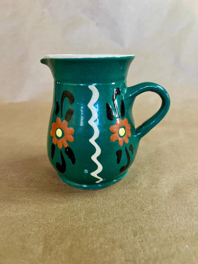 May include: A small, teal ceramic pitcher with a white rim and handle. The pitcher is decorated with orange and yellow floral designs and a white zigzag pattern. The pitcher is approximately 4 inches tall.