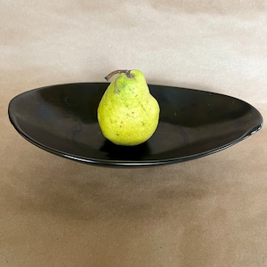 May include: A single green pear sits in the center of a black oval-shaped serving dish. The dish is on a brown paper background.