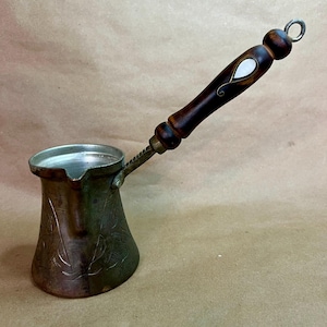 May include: A metallic coffee pot with a wide base and a narrow pouring spout. The body features etched line patterns. It has a long, dark brown wooden handle with a teardrop-shaped white inlay and a small metal ring at the very end for hanging.