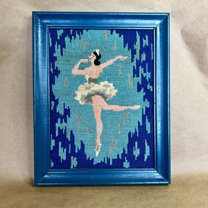 May include: A framed needlepoint artwork featuring a ballerina in a tutu. The artwork is set against a blue and turquoise background, framed in a blue wooden frame. The ballerina is depicted in shades of pink, white, and black.