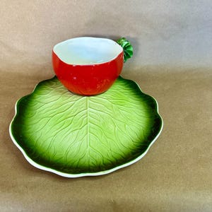 May include: A whimsical ceramic teacup and saucer set. The teacup is shaped like a red radish with a green stem handle, resting on a green leaf-shaped saucer. The set is designed with a bright, playful aesthetic, perfect for a unique kitchen collection.