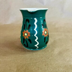 May include: A small, green ceramic pitcher with a white rim and spout. The pitcher is decorated with orange and black floral designs and a white wavy line. A charming, vintage-style creamer.