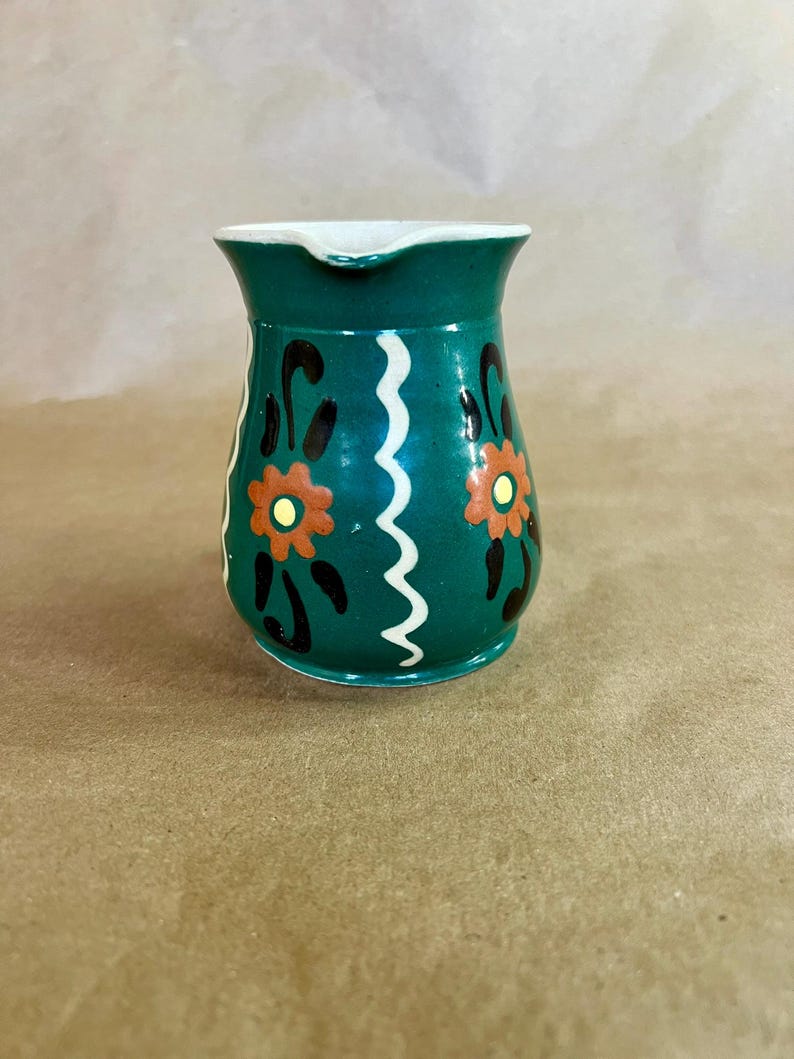 May include: A small, green ceramic pitcher with a white rim and spout. The pitcher is decorated with orange and black floral designs and a white wavy line. A charming, vintage-style creamer.