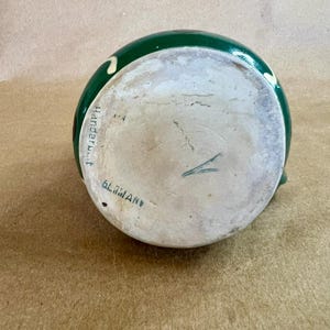 May include: A green ceramic pitcher with a white interior. The pitcher has a dark green exterior with white and yellow accents. The bottom of the pitcher has the text "GLIMAN" and "Handar.t."