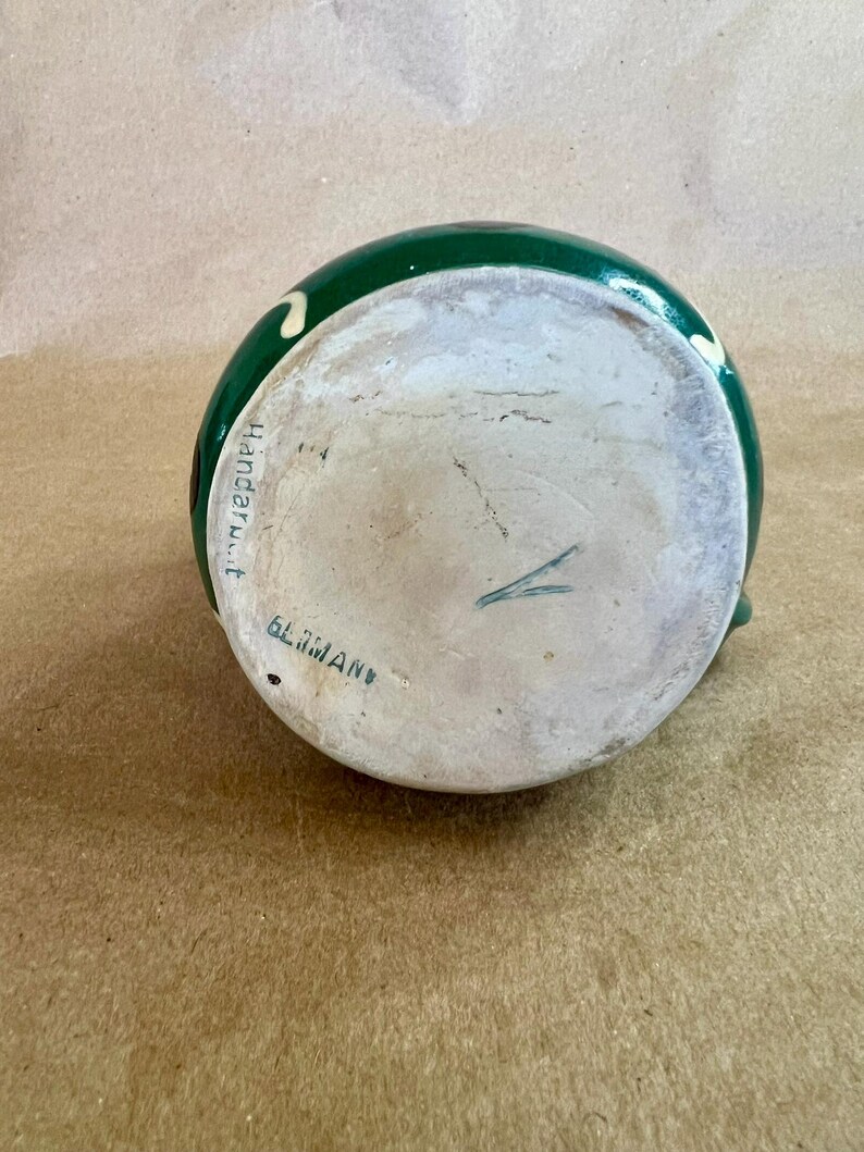 May include: A green ceramic pitcher with a white interior. The pitcher has a dark green exterior with white and yellow accents. The bottom of the pitcher has the text "GLIMAN" and "Handar.t."