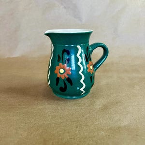 May include: A small, green ceramic pitcher with a handle and a spout. The pitcher is decorated with orange and black floral designs and white wavy lines. The interior is white.