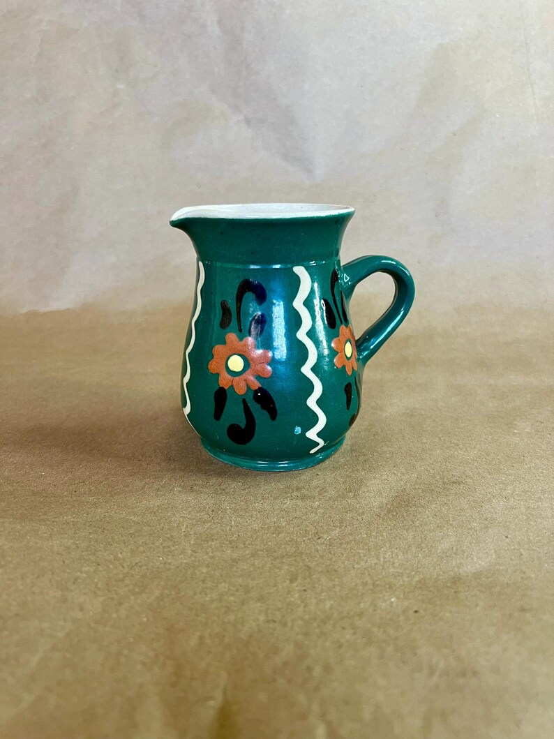 May include: A small, green ceramic pitcher with a handle and a spout. The pitcher is decorated with orange and black floral designs and white wavy lines. The interior is white.