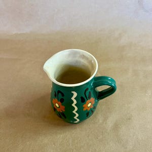 May include: A small, green ceramic pitcher with a handle and a spout. The pitcher is decorated with orange and black floral designs and a white zigzag pattern. The interior is off-white.