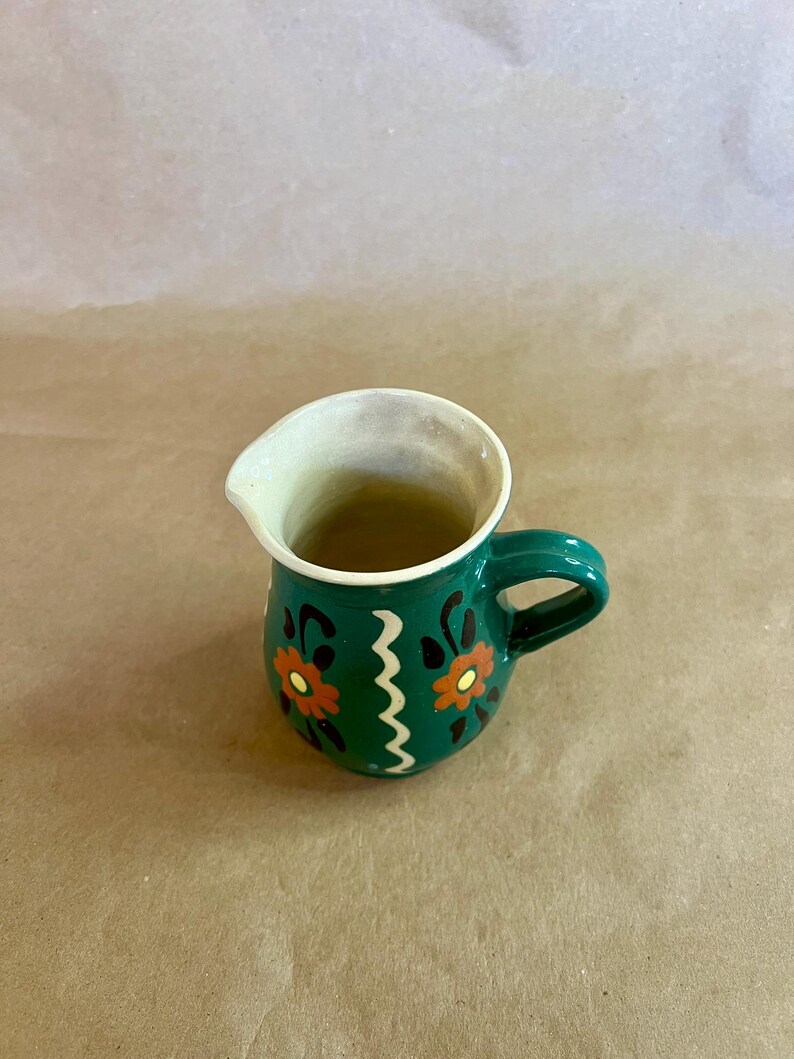 May include: A small, green ceramic pitcher with a handle and a spout. The pitcher is decorated with orange and black floral designs and a white zigzag pattern. The interior is off-white.