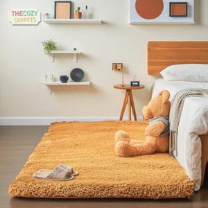 K&ouml;nnte beinhalten: Ein rechteckiger, goldgelber Teppich mit flauschiger Textur. Ein gro&szlig;er Teddyb&auml;r mit grauem Pullover sitzt auf dem Teppich in der N&auml;he eines Bettes. Das Cozy Carpets-Logo ist sichtbar. Hausschuhe liegen auf dem Teppich.
