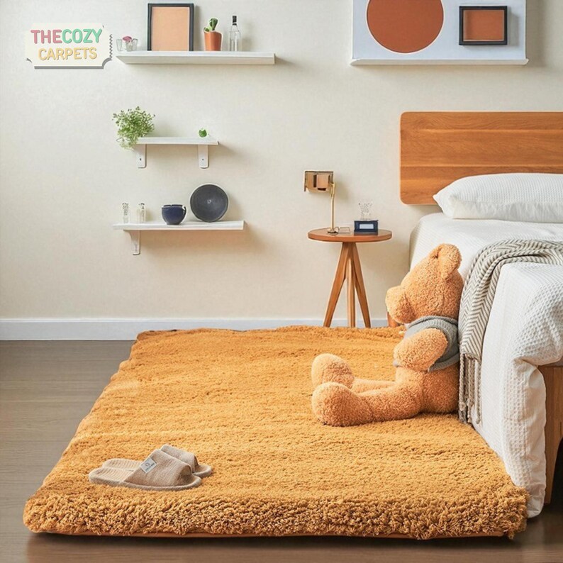 K&ouml;nnte beinhalten: Ein rechteckiger, goldgelber Teppich mit flauschiger Textur. Ein gro&szlig;er Teddyb&auml;r mit grauem Pullover sitzt auf dem Teppich in der N&auml;he eines Bettes. Das Cozy Carpets-Logo ist sichtbar. Hausschuhe liegen auf dem Teppich.