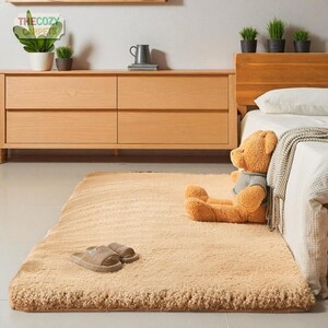 K&ouml;nnte beinhalten: Ein rechteckiger, beigefarbener, flauschiger Teppich mit zotteliger Textur. Ein Paar braune Hausschuhe und ein Teddyb&auml;r mit grauer Weste liegen auf dem Teppich. Der Teppich befindet sich in einem Schlafzimmer, in der N&auml;he einer Holzkommode und eines Bettes.