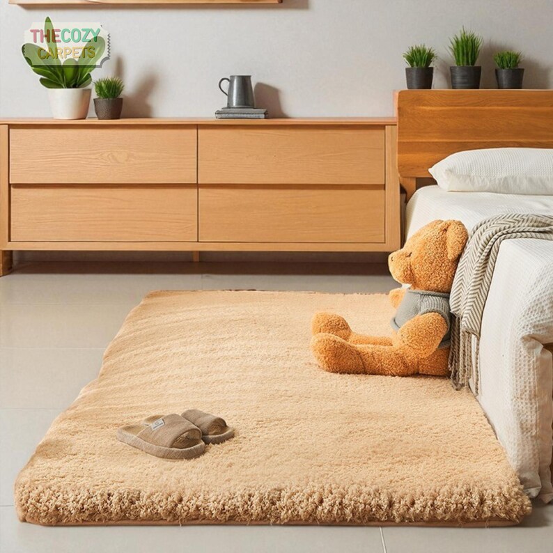 K&ouml;nnte beinhalten: Ein rechteckiger, beigefarbener, flauschiger Teppich mit zotteliger Textur. Ein Paar braune Hausschuhe und ein Teddyb&auml;r mit grauer Weste liegen auf dem Teppich. Der Teppich befindet sich in einem Schlafzimmer, in der N&auml;he einer Holzkommode und eines Bettes.