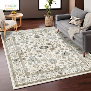 Pode incluir: Um tapete retangular com um intrincado padrão floral em tons de creme, cinza e azul suave. O tapete apresenta uma borda detalhada e está colocado sobre um chão de madeira escura. O logotipo The Cozy Carpets é visível no canto superior esquerdo.