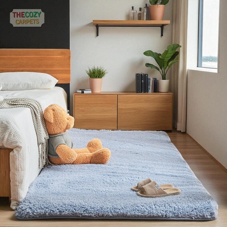K&ouml;nnte beinhalten: Ein hellblauer, rechteckiger, flauschiger Teppich in einem Schlafzimmer. Ein Teddyb&auml;r sitzt auf dem Teppich in der N&auml;he eines Bettes. Der Teppich ist etwa 152 cm lang und 102 cm breit. Das Cozy Carpets-Logo befindet sich oben links.