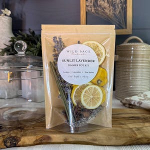 May include: A clear, resealable pouch containing a Sunlit Lavender simmer pot kit. The label reads Wild Sage Handmade, Sunlit Lavender, Simmer Pot Kit, Lemon, Lavender, Star Anise, Fresh bright & calming. Dried lemon slices, lavender, and star anise are visible.