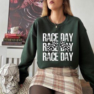 May include: A dark green sweatshirt with the text "RACE DAY" repeated three times. The text is in white and has a distressed look. Two black and white checkered flags are crossed in the middle of the text.
