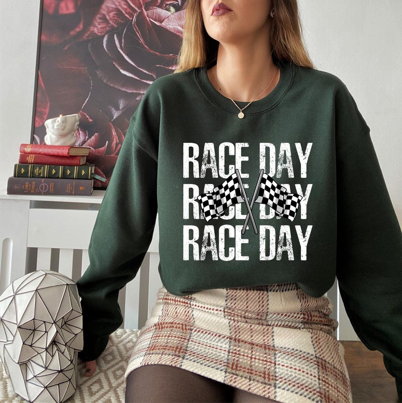 May include: A dark green sweatshirt with the text "RACE DAY" repeated three times. The text is in white and has a distressed look. Two black and white checkered flags are crossed in the middle of the text.