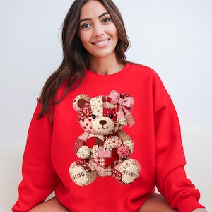 May include: Red sweatshirt featuring a patchwork teddy bear design. The bear has a pink bow, a heart, and the words "Love", "Hugs", and "Kisses". The sweatshirt is a solid red color.