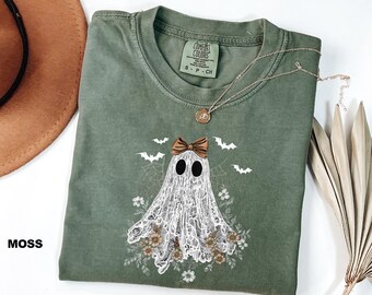 May include: A moss green t-shirt with a ghost design. The ghost has a brown bow, black eyes, and a lace-like body. Bats, flowers, and spiderwebs surround the ghost. The word "MOSS" is printed on the bottom left of the t-shirt.
