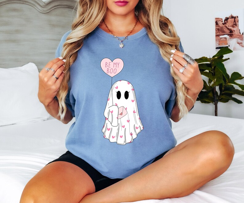 May include: A light blue t-shirt with a ghost holding a pink heart balloon that says "Be My Boo". The ghost is covered in pink hearts.
